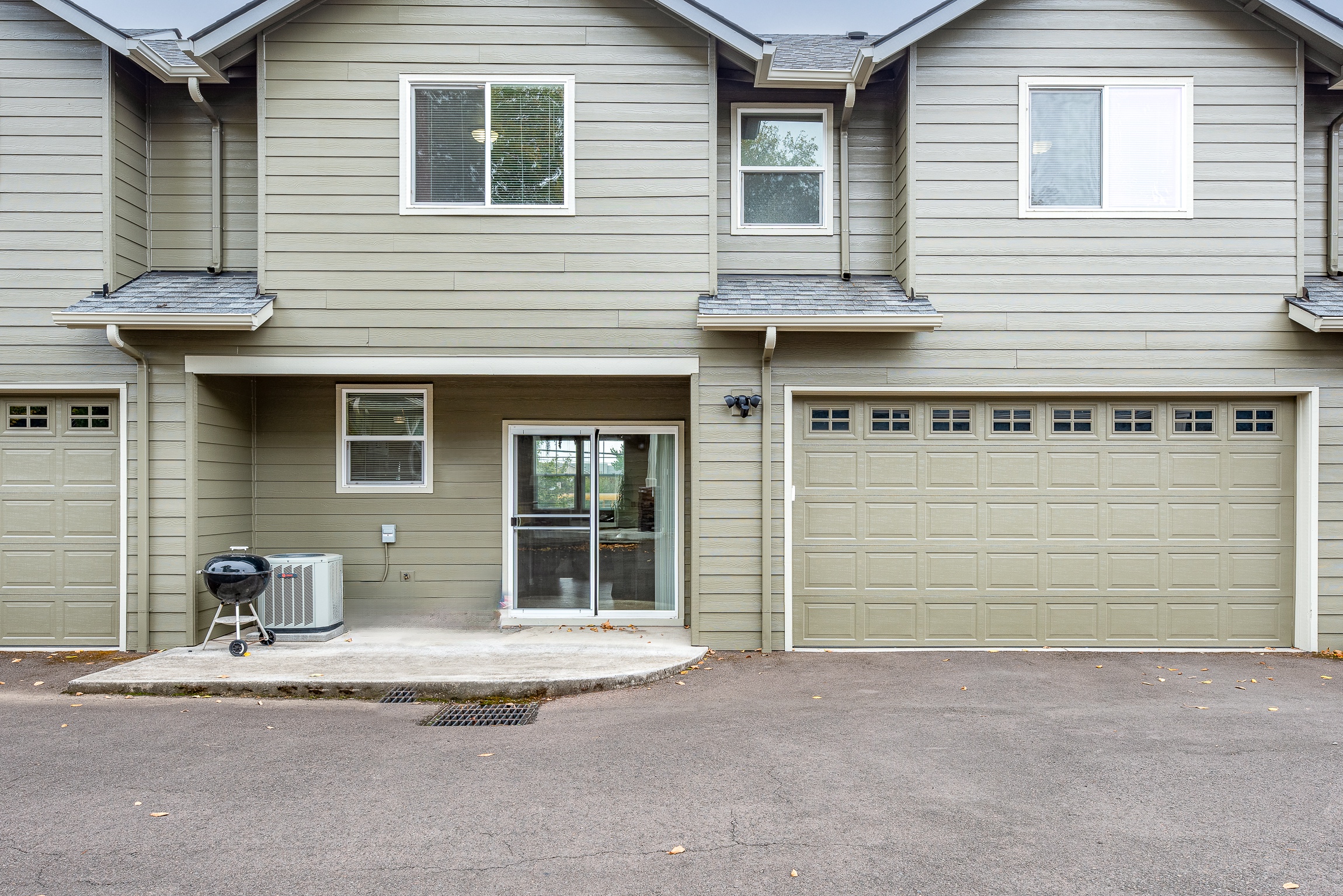 A house exterior featuring beige siding, a sliding glass door, a large garage door with windows, an air conditioning unit, and a small charcoal grill on a concrete patio, all set against a paved driveway.