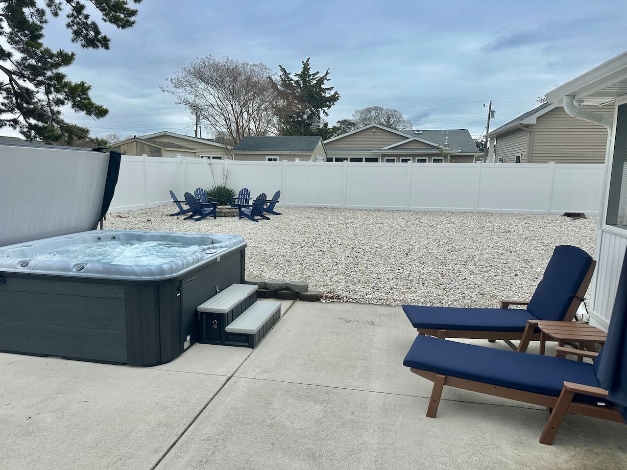 Unwind in your private outdoor retreat featuring a bubbling hot tub and comfortable lounge chairs with navy cushions. The spacious, fenced-in yard also includes a gravel area with Adirondack chairs gathered around a fire pit, perfect for starlit evenings.