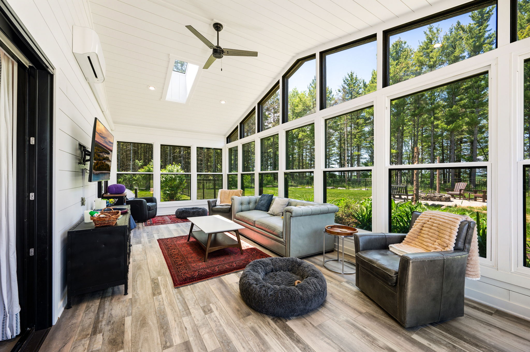 Kick back in the spacious sunroom lounge, featuring a comfy leather sofa, mounted smart-TV, and panoramic windows overlooking the woods.
