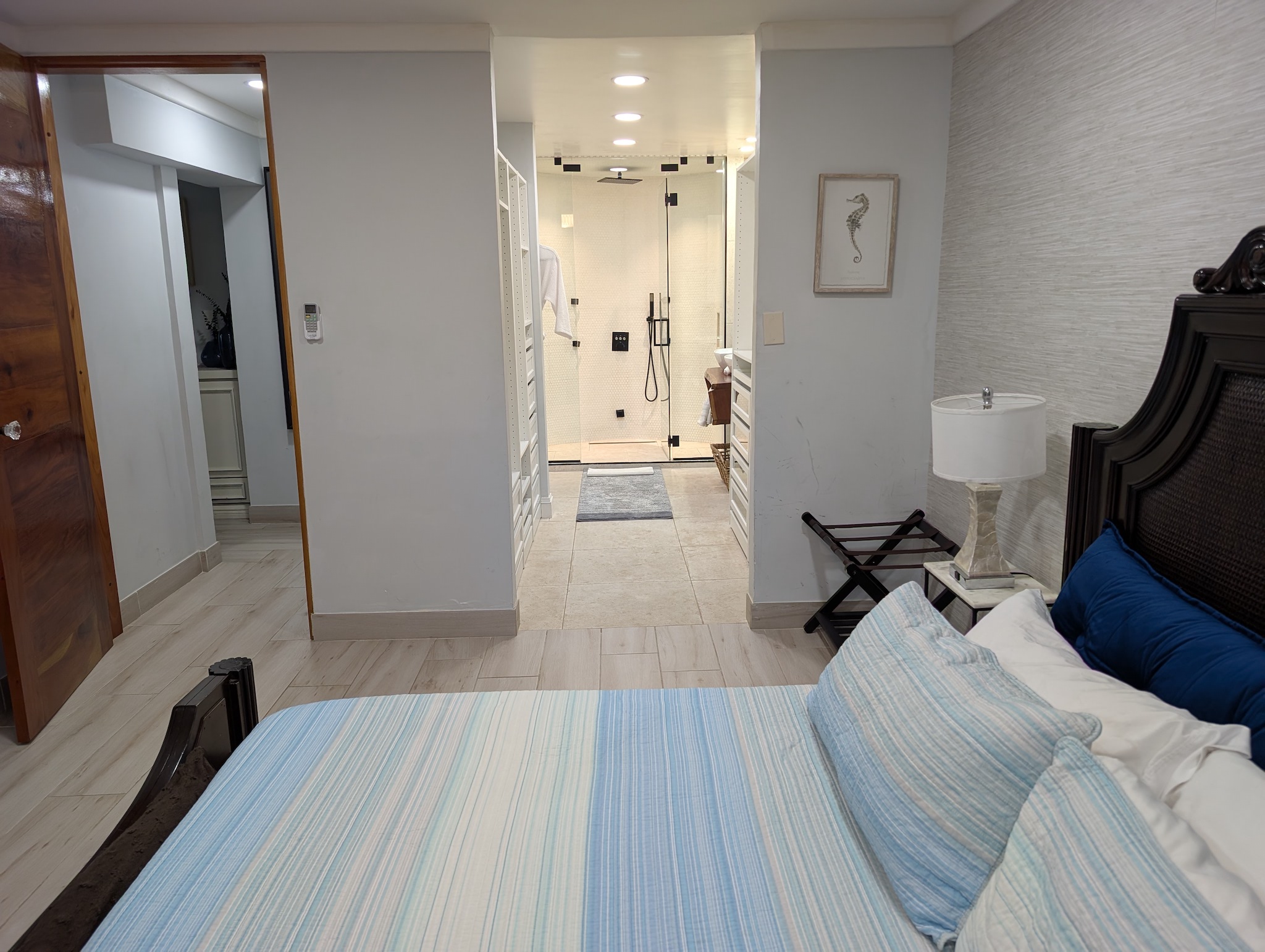 Primary bedroom at Villa Coral featuring a dark wood king bed frame, coastal blue bedding, and a direct view into the walk-in closet and glass steam shower in Dorado, Puerto Rico.
