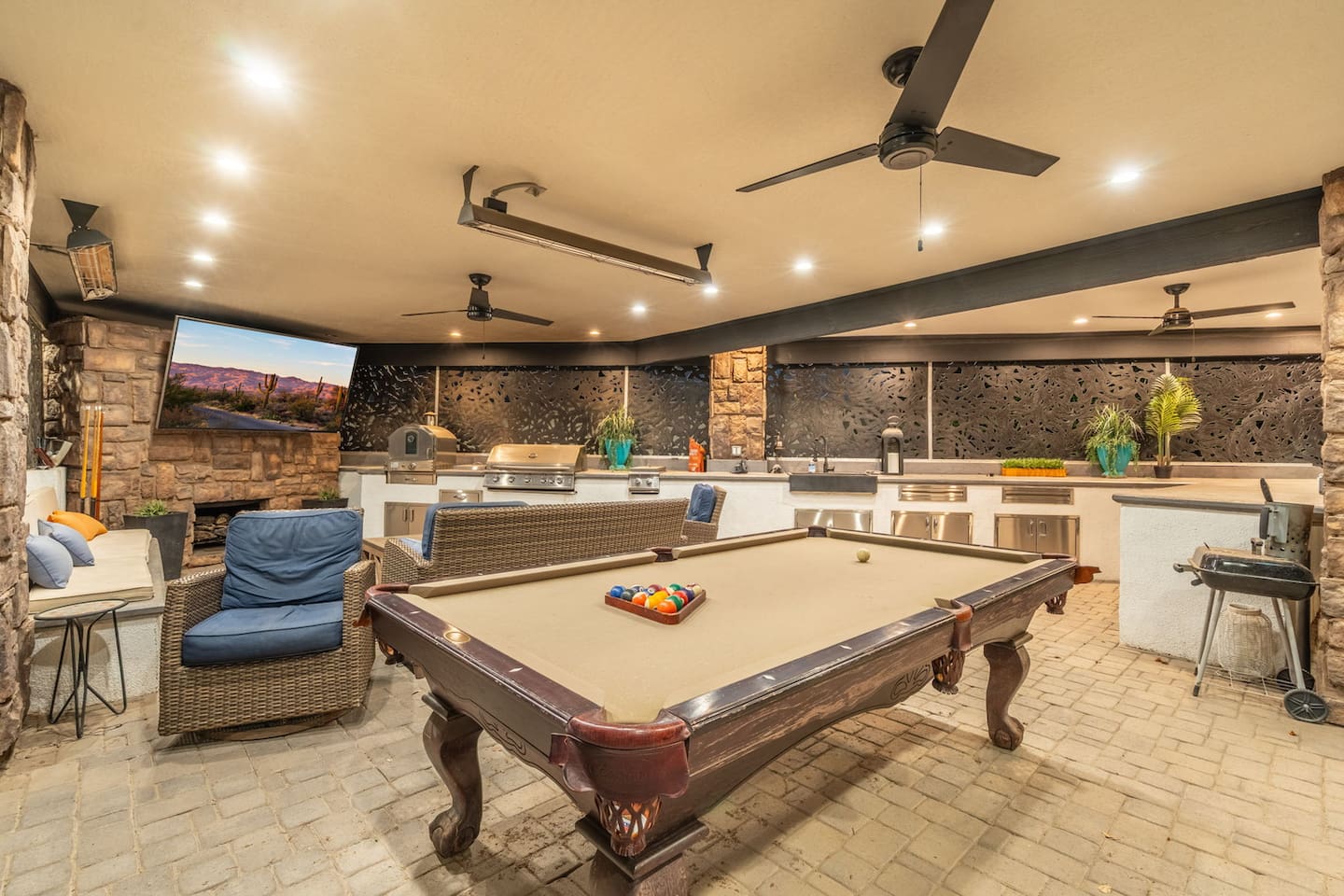  Covered outdoor lounge with a pool table, built-in kitchen, cozy fireplace seating, and mounted TV — an entertainer's dream under the desert stone accent wall.