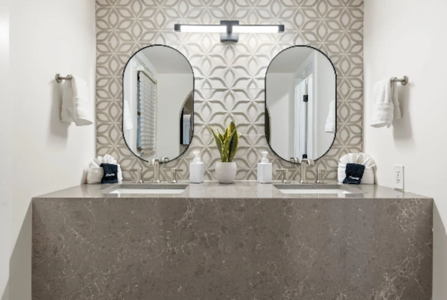 A dual vanity in patterned stone tile sets a spa-like tone, with a moody black-tiled shower just beyond. Every surface here feels intentional, right down to the oval mirrors and brushed fixtures.