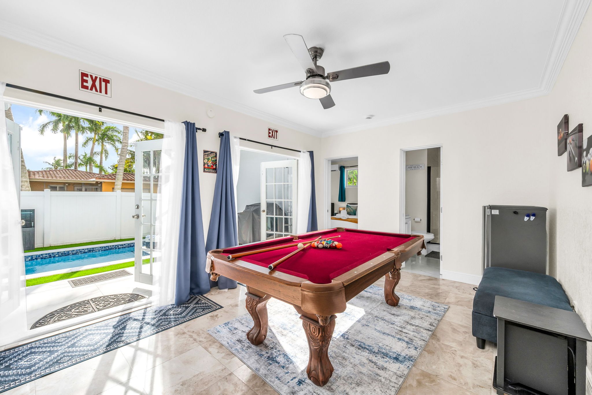 Bright Florida sunroom with a full-size pool table, seating area, beverage cooler, and a streaming TV. French doors open directly to the pool deck for seamless indoor–outdoor entertainment.
