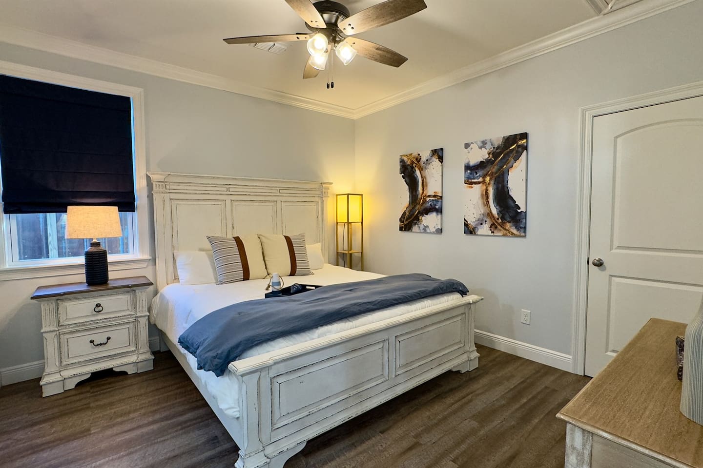 A stylish, light-filled bedroom showcasing a timeless white bed frame, modern artwork, warm wood flooring, and a serene layout designed for restful nights.