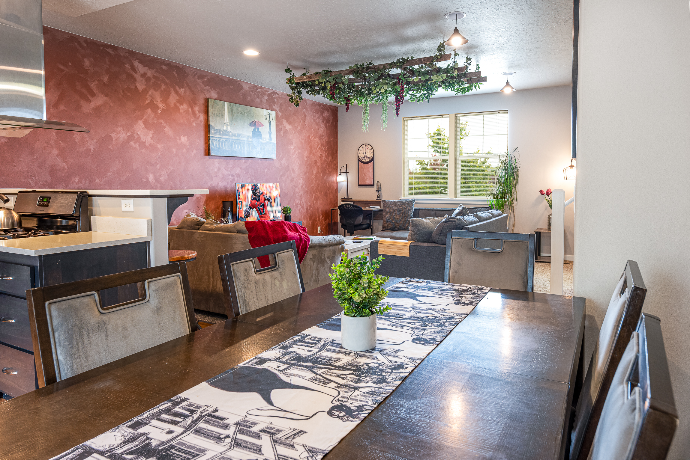 A modern, open-concept living space featuring a dark wooden dining table with a decorative runner, a stainless steel stove in the kitchen area, a cozy seating arrangement with a grey couch and an armchair, a wall adorned with a clock and artwork, and greenery suspended from the ceiling, with large windows allowing natural light to flow in.