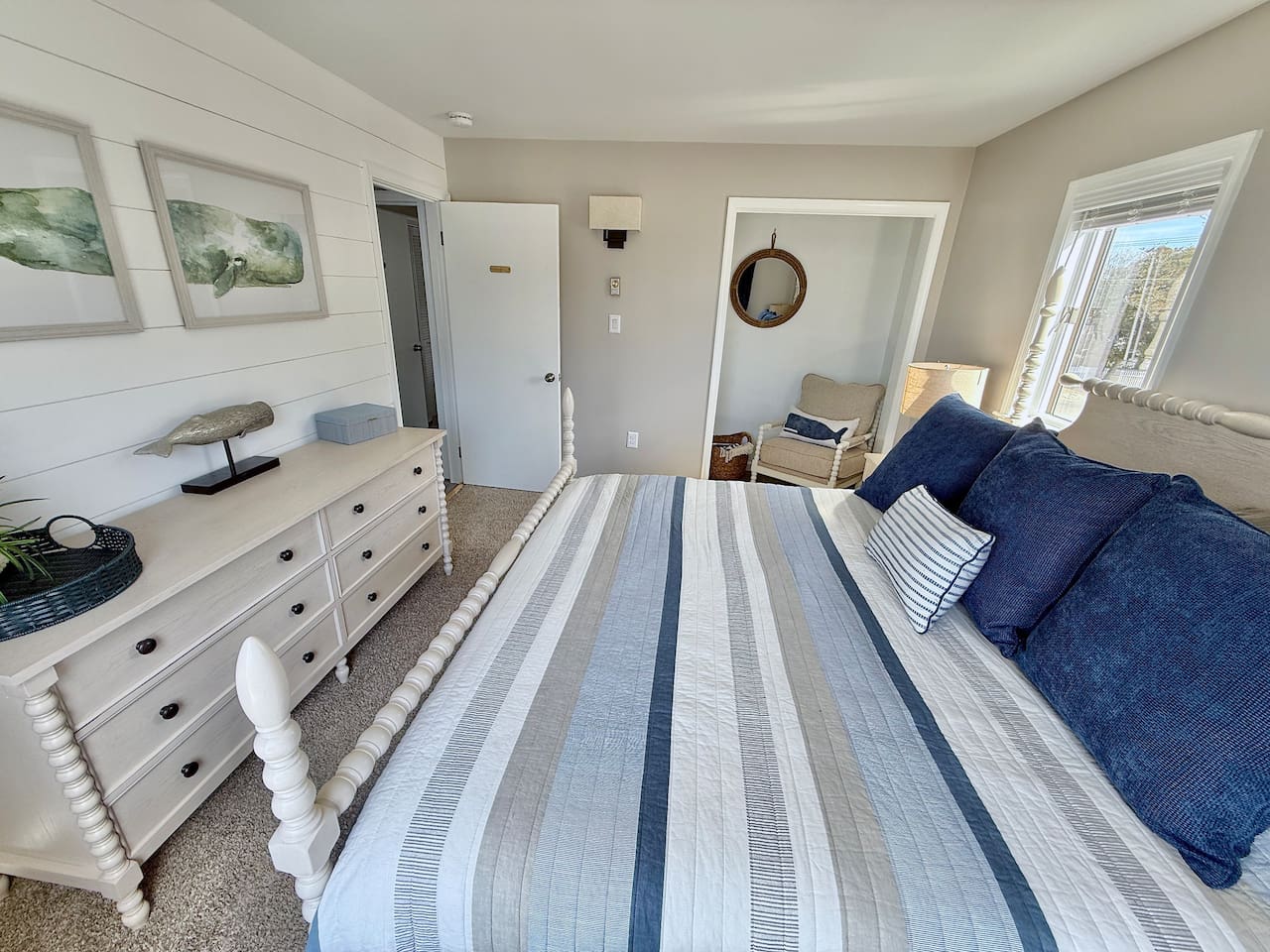 This coastal-themed bedroom features a white spindle bed with blue-striped bedding, a matching dresser with whale-themed decor, and a cozy reading nook, all coming together to create a relaxing, nautical-inspired escape.