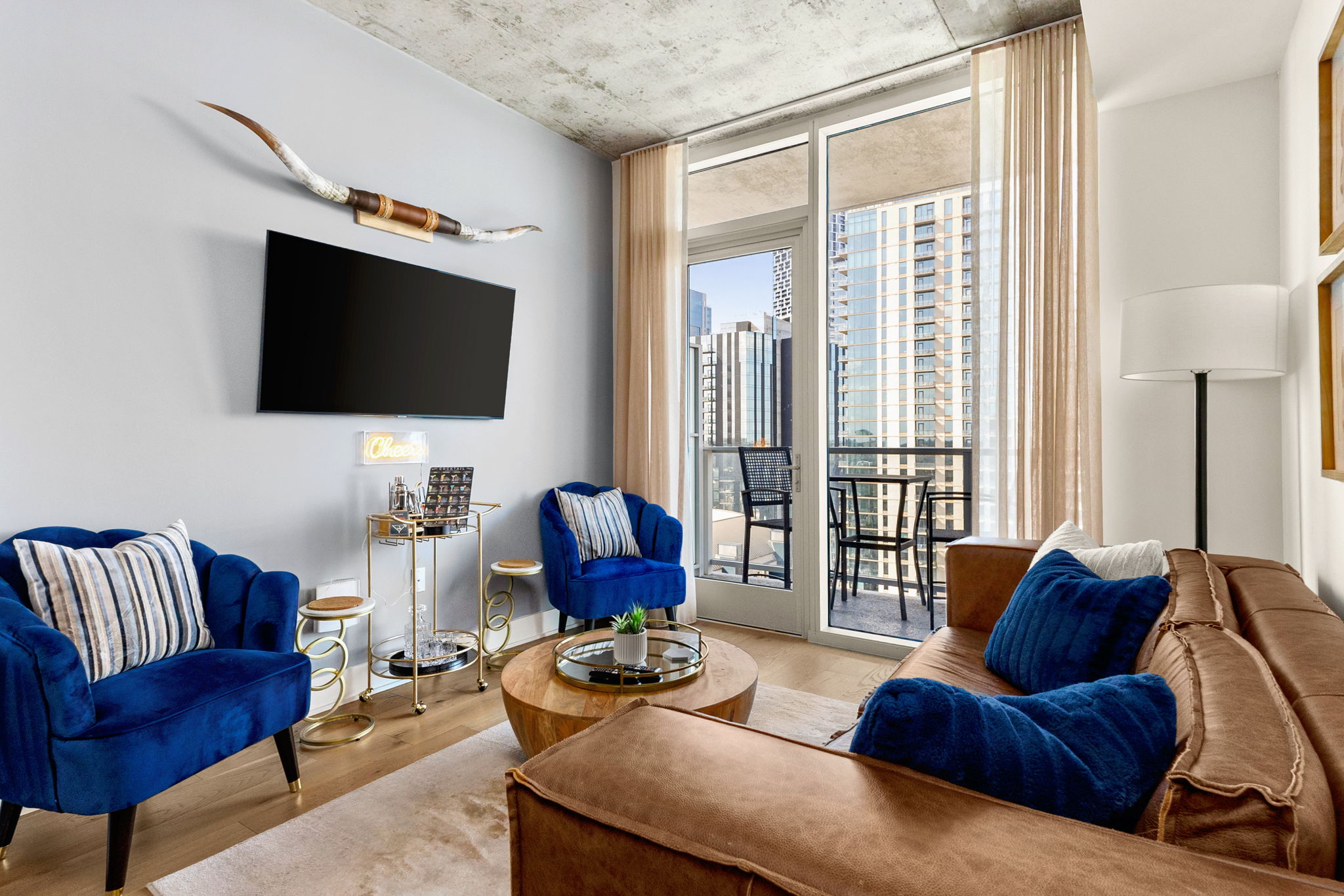 A vibrant living room featuring a brown leather sofa and two striking blue armchairs, with a large glass door leading to a balcony that offers a view of a modern cityscape.