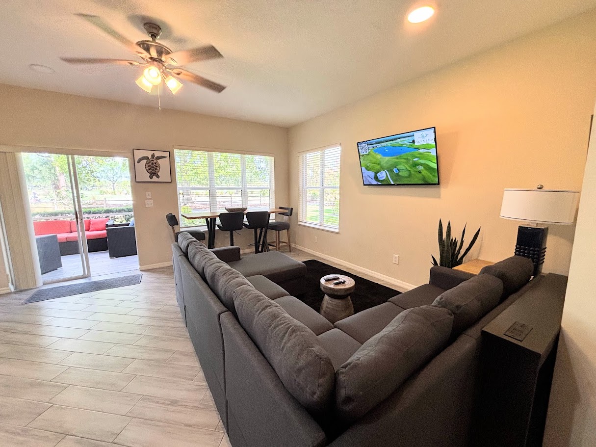 Spacious living and dining area with a large sectional, wall-mounted TV, and natural light from the patio doors.