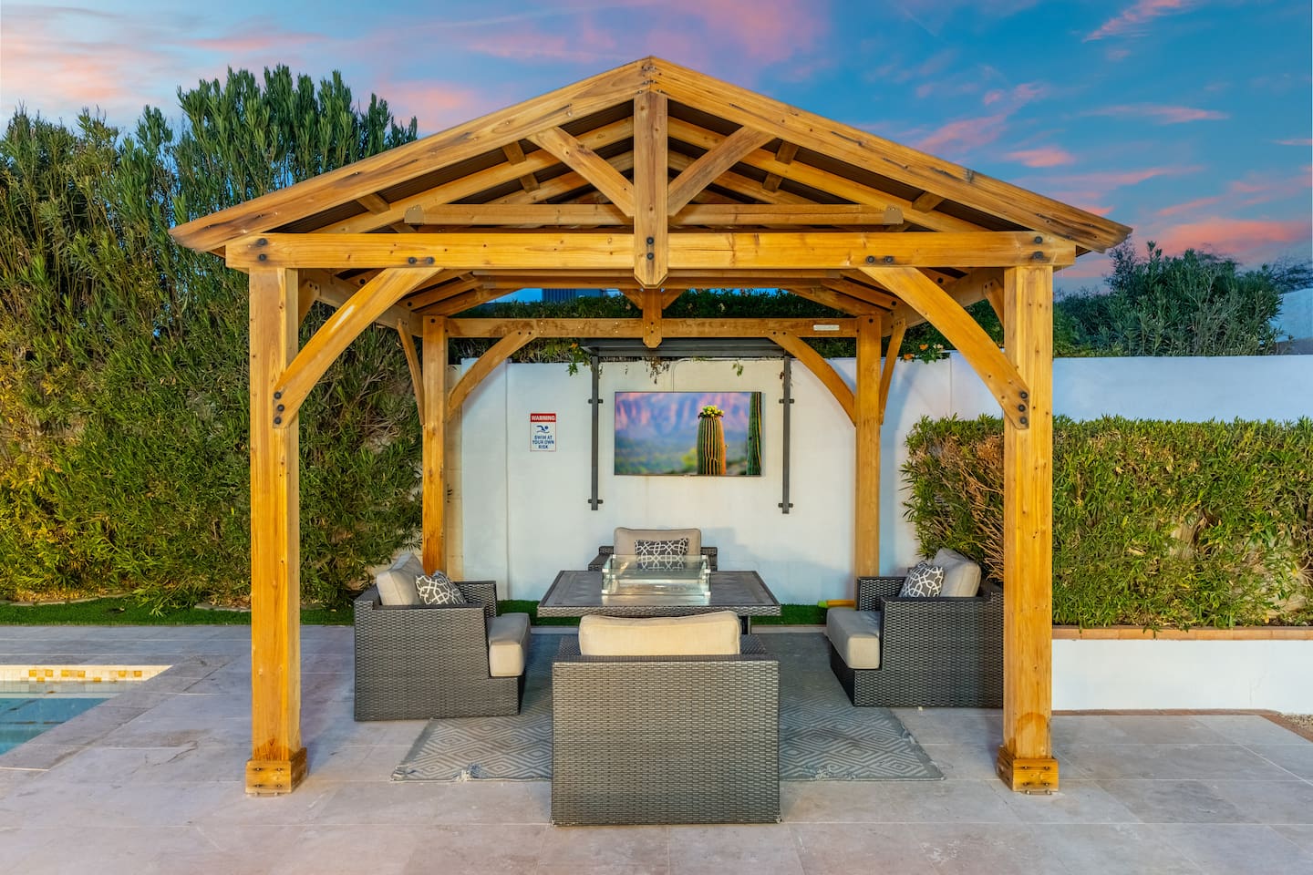 The wooden pergola shelters a warm fire table and stylish wicker seating—ideal for intimate conversations or movie nights on the outdoor TV.