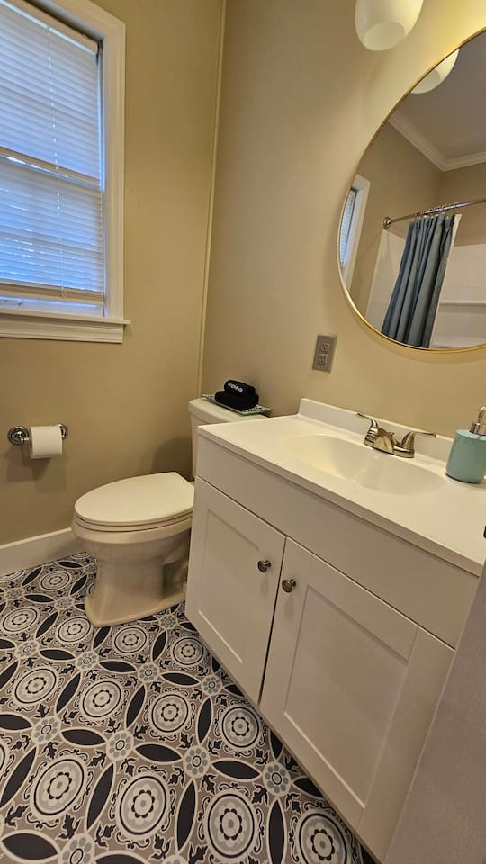 Charming bathroom with a sleek white vanity, bold patterned tile flooring, and a large round mirror for a fresh, modern feel.