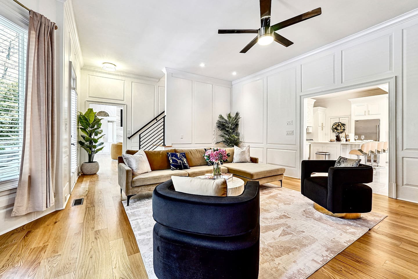 Situated next to the kitchen, this main-floor living room offers a warm, upscale ambiance with a brown L-shaped sectional, two black accent chairs, gold and black design accents, a smart TV, and a fireplace, perfect for entertaining or unwinding.
