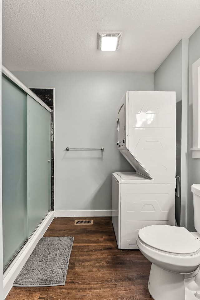 Full bathroom view showing the glass-enclosed shower, stacked washer and dryer, and durable hardwood-style flooring.