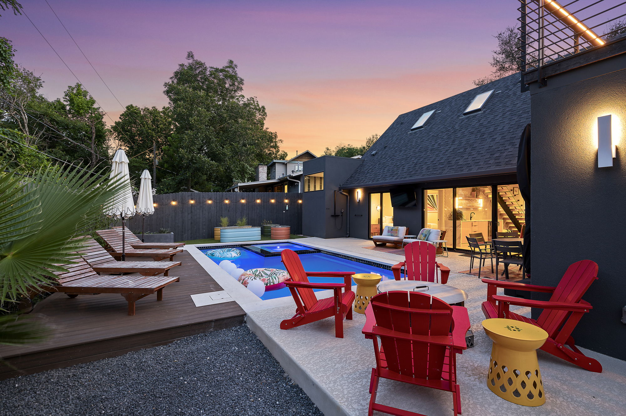 A stunning backyard oasis is illuminated at sunset, showcasing a sparkling swimming pool with a hot tub and a multi-level deck for dining and lounging.