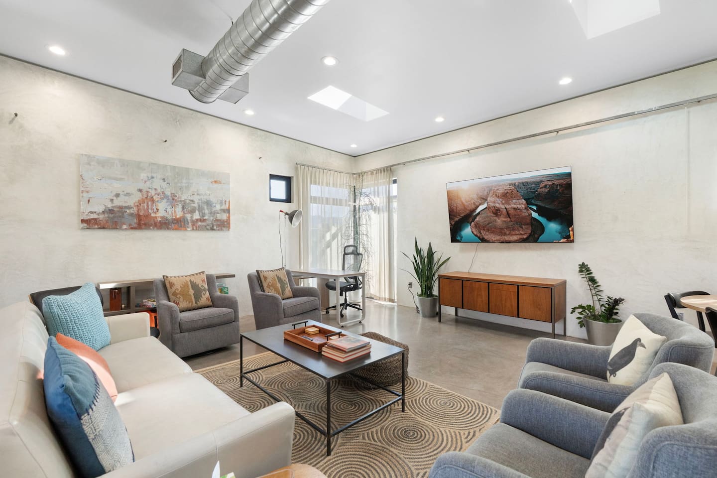 Spacious modern living room with comfortable seating, a mounted TV, workspace nook, and plenty of natural light.