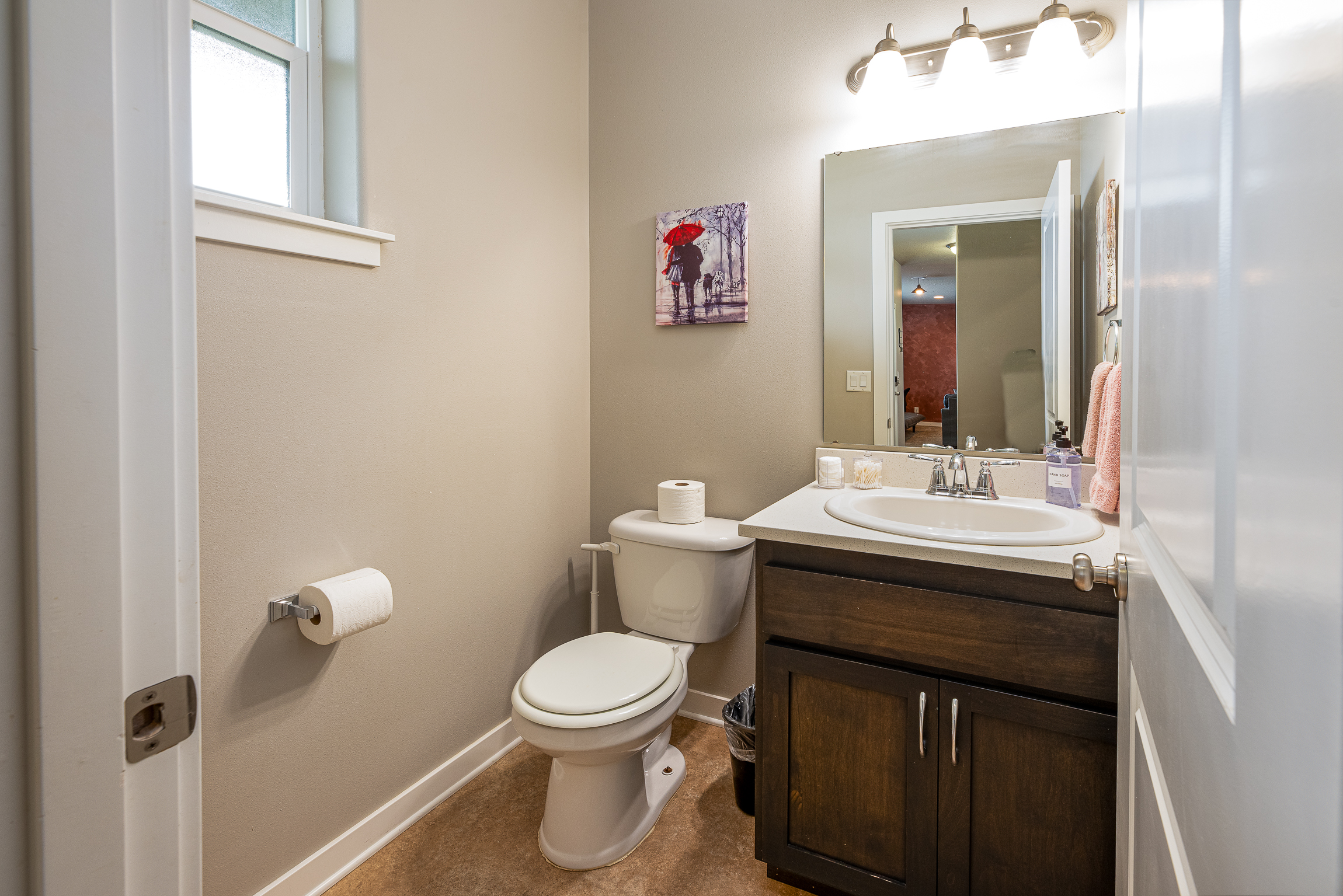 A small bathroom featuring a white toilet, a sink with dark wood cabinetry, a mirror above the sink, a roll of toilet paper on the wall, a light fixture with three bulb lights, and a artwork of a couple with an umbrella hanging on the wall above the toilet.