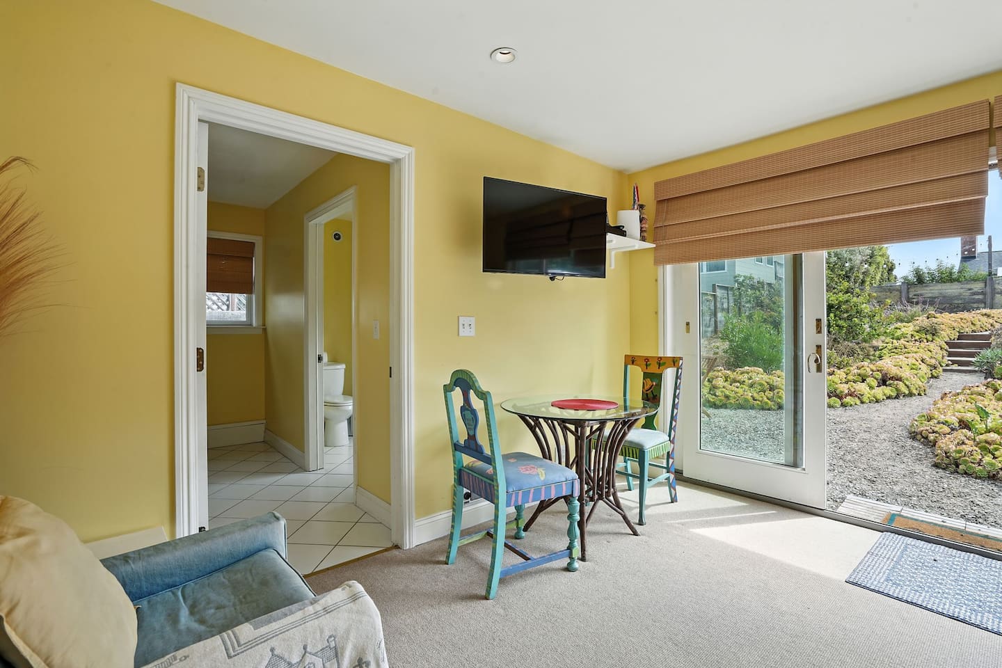 - Bright sitting area with French doors opening to the garden and patio seating  
- Includes a small dining set, wall mounted TV, and easy access to the bathroom  
- Cheerful yellow walls create a warm and inviting space for guests to enjoy  