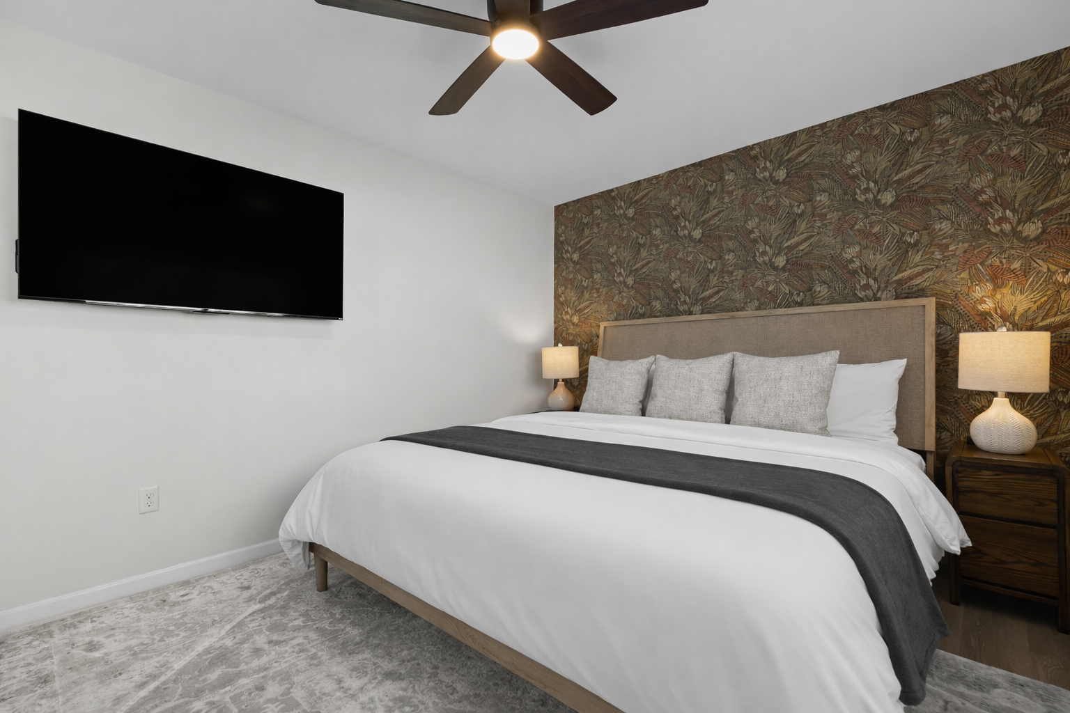 Rest easy in this modern bedroom with a plush bed, stylish accent wall, smart TV, and ceiling fan—perfect for cozy nights and relaxed mornings.