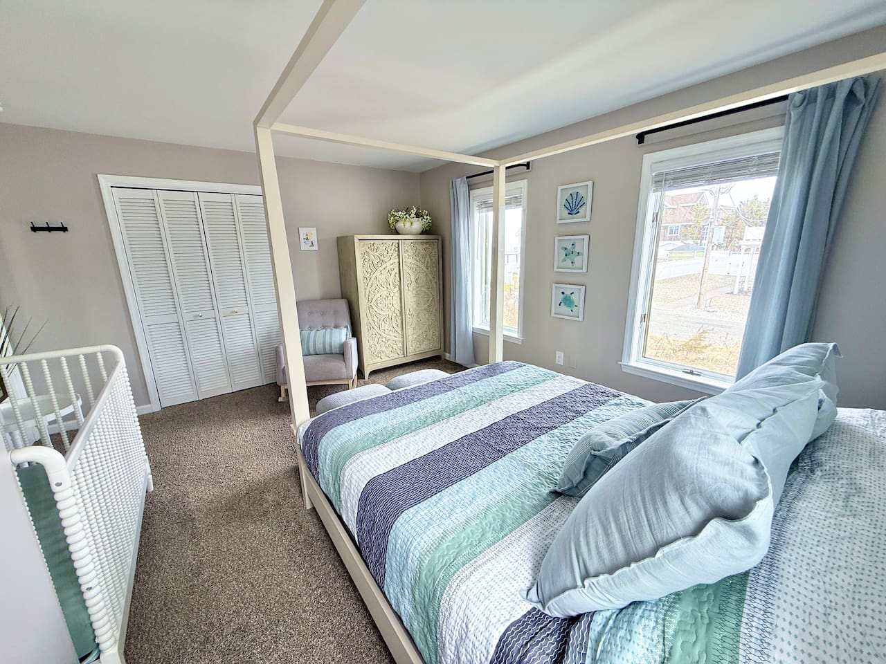 This family-friendly bedroom features a four-poster canopy bed with striped bedding and a white crib. A cozy rocking chair, decorative armoire, and coastal-themed wall art complete the serene space.