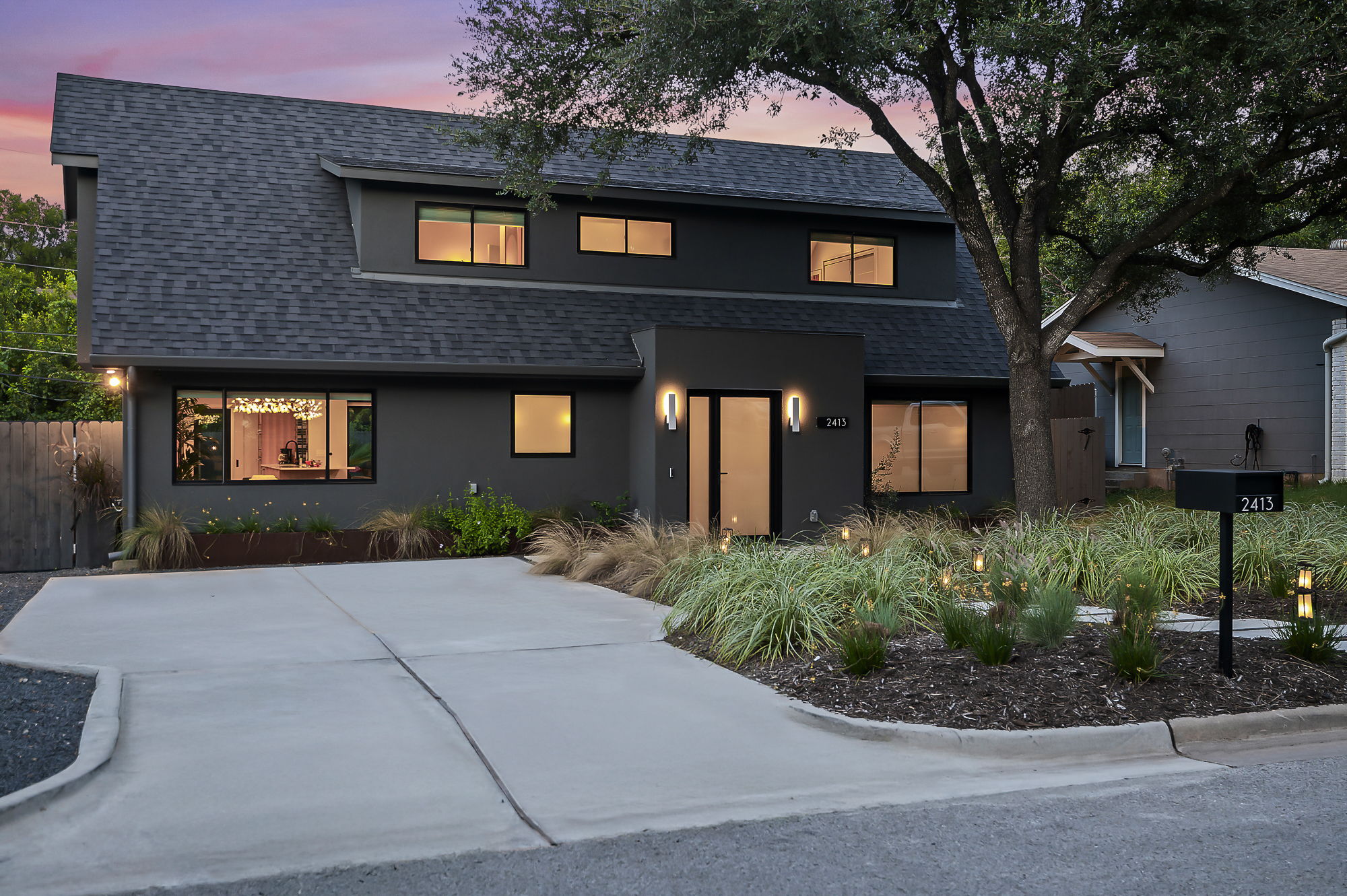 A contemporary dark gray home with a multi-level roofline, a minimalist-style front yard with a concrete driveway, and modern exterior lighting, all captured at dusk with a hint of color in the sky.