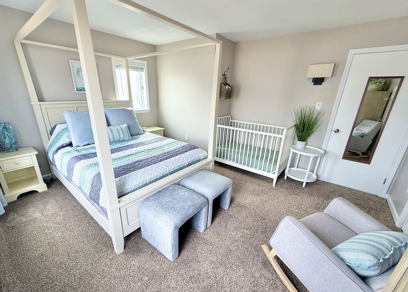 This family-oriented bedroom offers a serene retreat with a four-poster canopy bed, a white crib, and a cozy gray rocking chair. The space is completed with stylish ottomans, a side table with greenery, and a mirror.