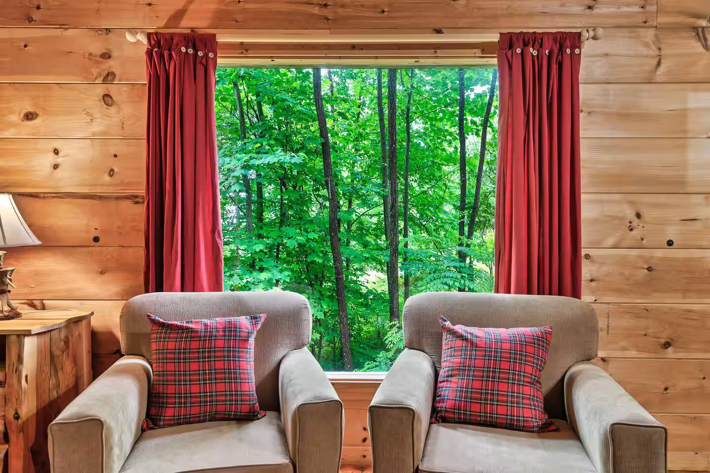 A serene seating area with plush chairs and cheerful plaid accents is complemented by a window framing the vibrant greenery, perfect for sipping coffee and enjoying the natural surroundings.