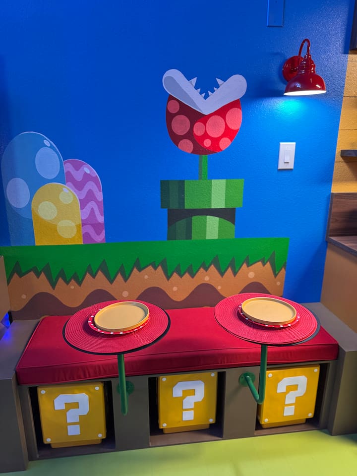 Family-friendly Mario breakfast nook at Mario's Mini Mansion in Clermont, FL! Red cushioned bench, question mark block stools (removable), and giant Piranha Plant mural create a fun spot for pancakes and morning smiles.