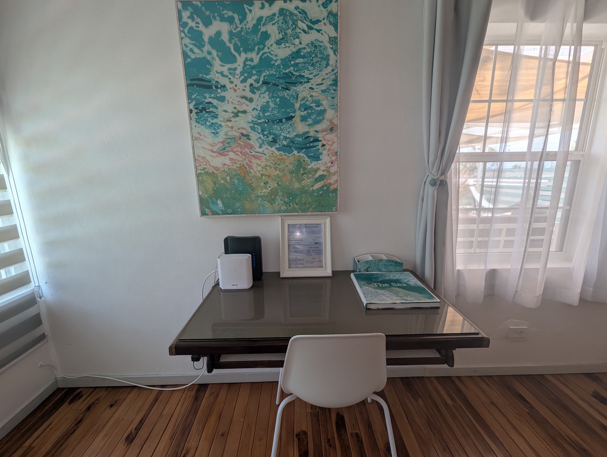 Dedicated remote workspace at Villa Coral featuring a glass-top desk, white modern chair, high-speed Wi-Fi router mesh pod, coastal wave art, and natural window lighting in Dorado, Puerto Rico.
