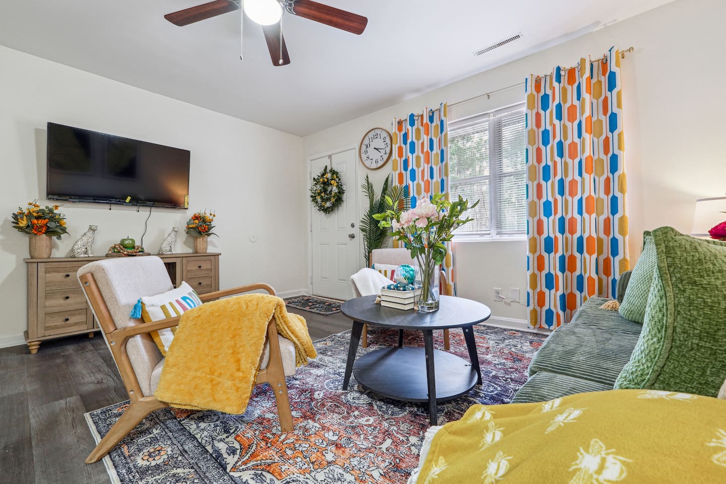 Enjoy a cozy living room featuring a long comfortable sofa, a TV for your entertainment, a ceiling fan, two chair, and a window with a lovely view outside — perfect for relaxing and unwinding.
