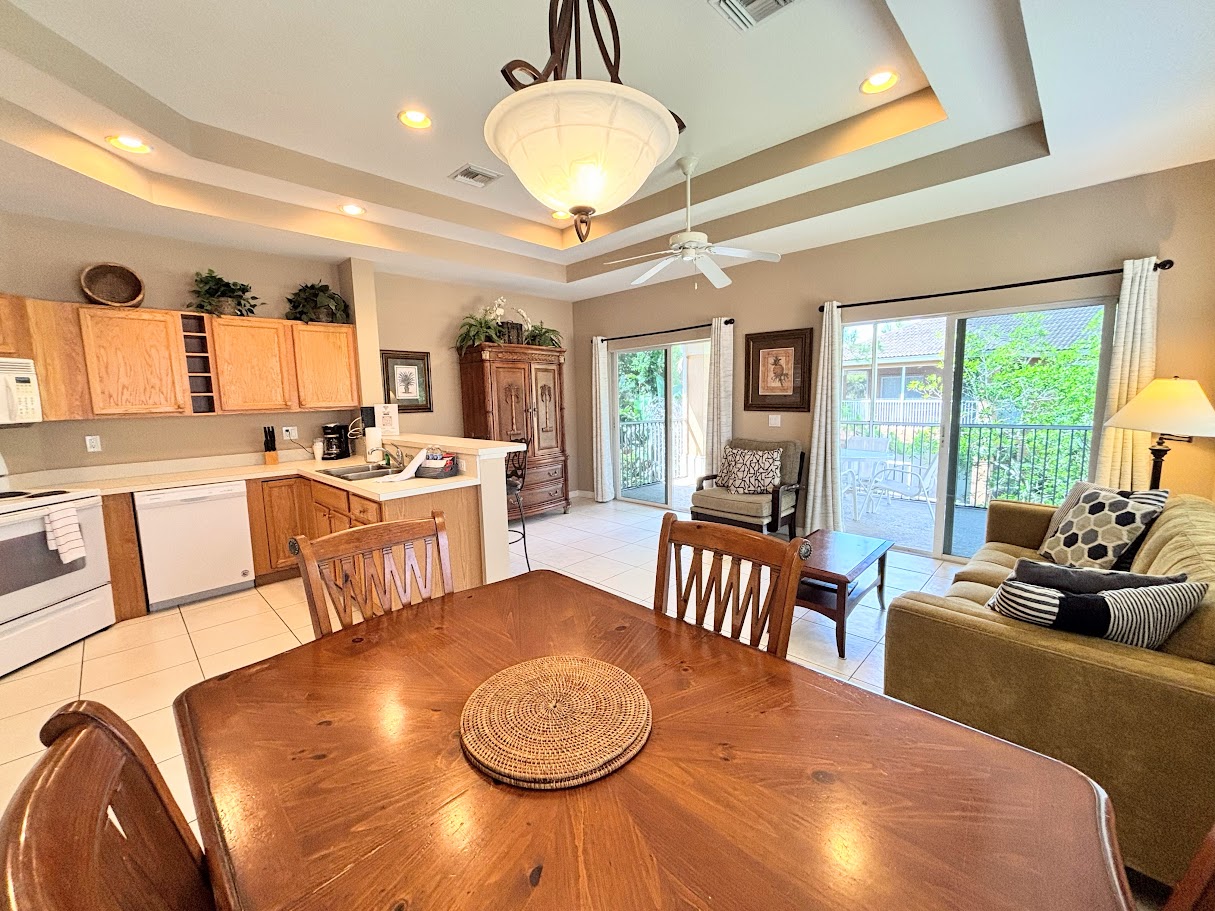 Bright and spacious open-concept living area featuring vaulted tray ceilings, a fully equipped kitchen, comfortable seating, and sliding glass doors leading out to a private screened balcony.