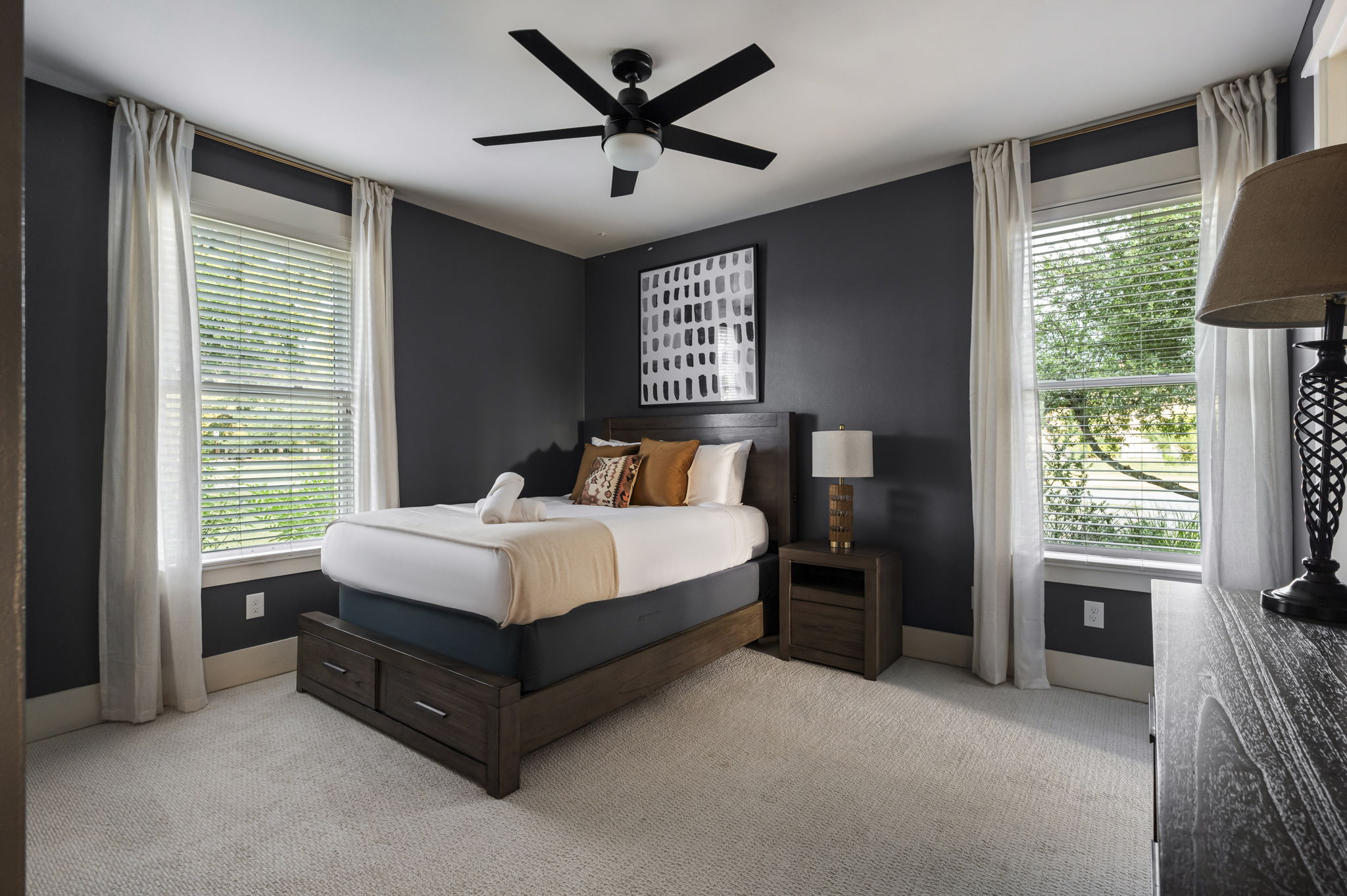Bright and cozy bedroom with a queen bed, ceiling fan, and two sunlit windows.