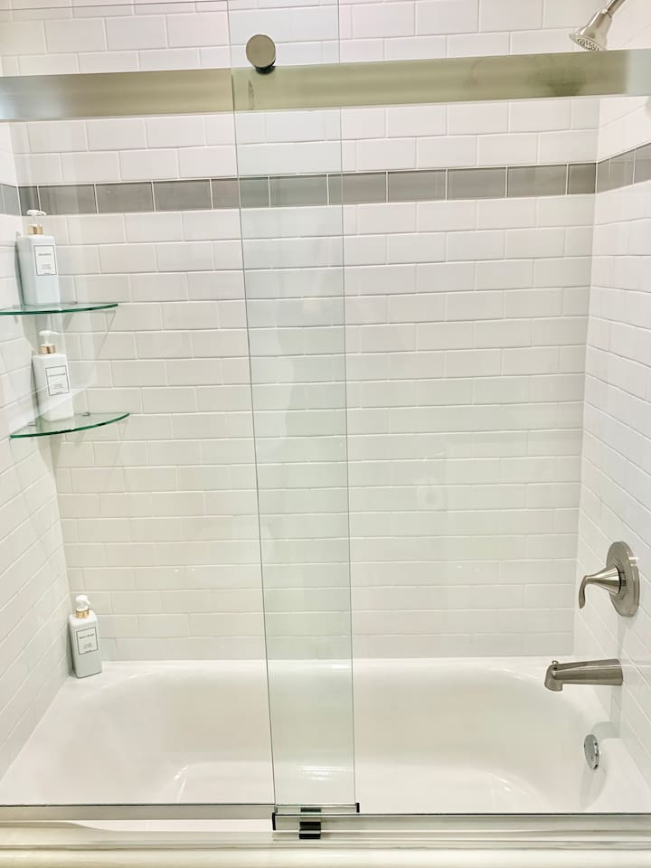 Enjoy a relaxing soak in the clean, white bathtub featuring modern brushed nickel fixtures. The space is beautifully finished with classic white subway tile and a sleek glass door, providing a bright and polished feel.
