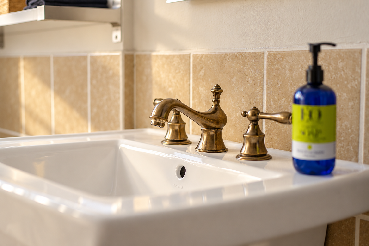 Bathroom detail featuring a bright white basin, traditional brass-toned taps and a warm beige tile backsplash. Sunlight picks out the curved faucet and clean-lined sink, offering a closer look at the bathroom finishes.