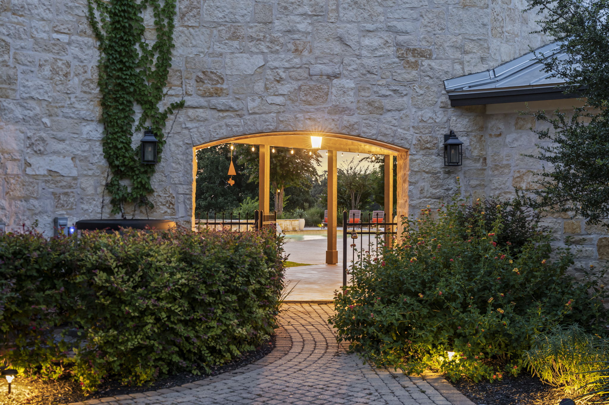 A welcoming stone archway leads to an outdoor area, framed by lush green bushes and illuminated by warm evening lights.