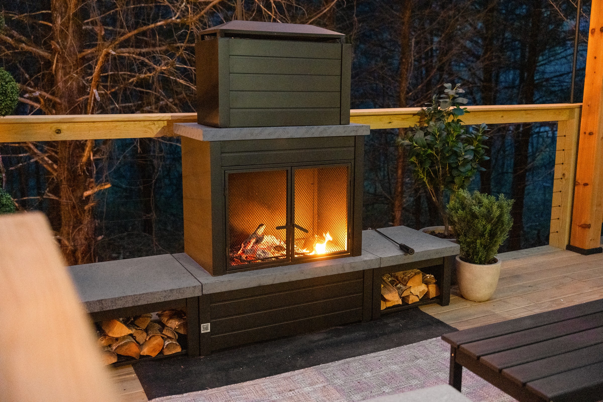 Wood-burning fireplace lit at night on the deck. Stacked firewood beside the flames makes it easy to keep the evening going.