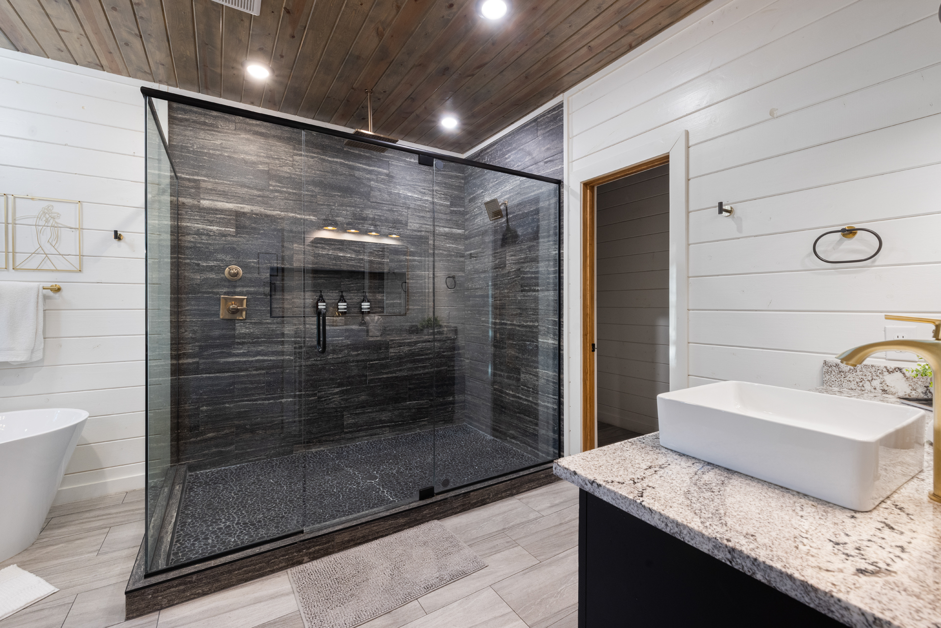 Enjoy a luxurious oversized walk-in shower featuring floor-to-ceiling charcoal stone tile, a pebble-style floor, rainfall showerhead, bronze fixtures, and frameless glass walls for a true spa-like retreat.