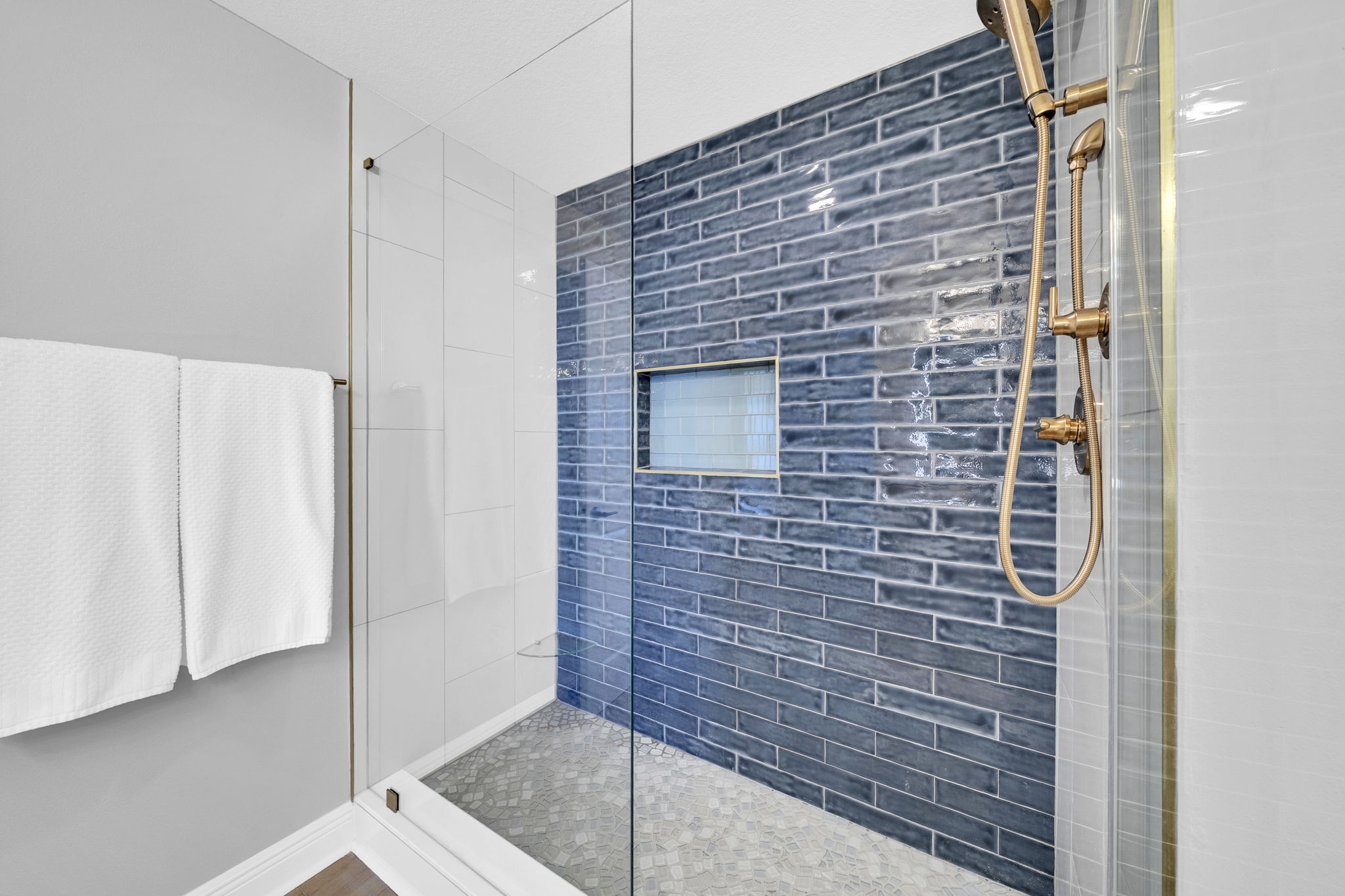 Step into a spa-like shower with stunning blue tile that mirrors the Gulf and sea glass–inspired flooring that brings the beach inside—complete with a handheld sprayer and a sleek glass toe ledge for added comfort.