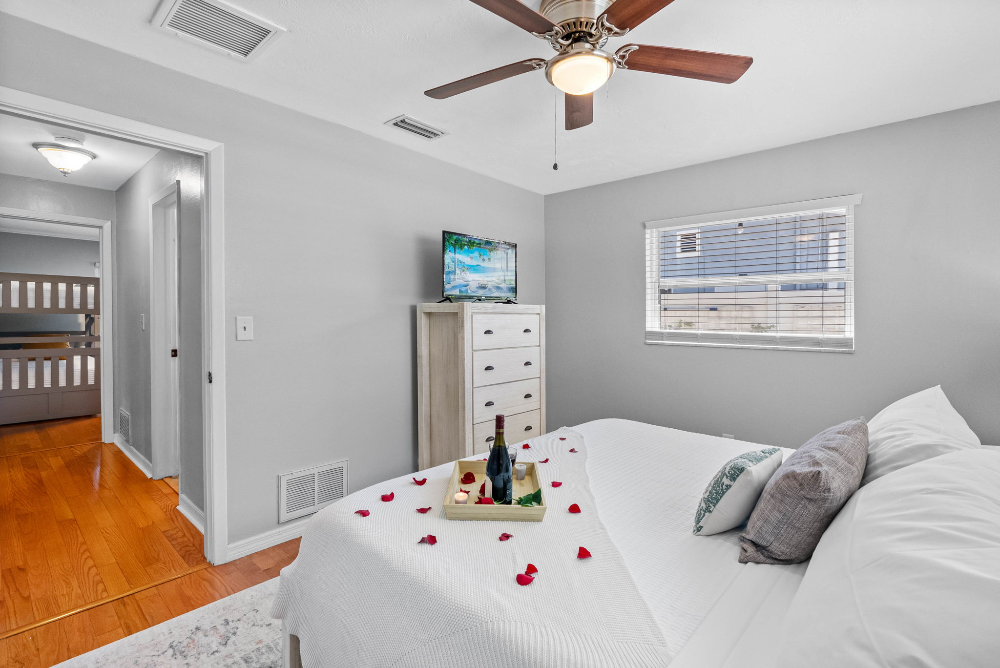 Relax in this peaceful bedroom featuring a crisp white bedspread, soft accent pillows, and subtle beachy decor. A curated tray with wine and candles adds a touch of indulgent charm.