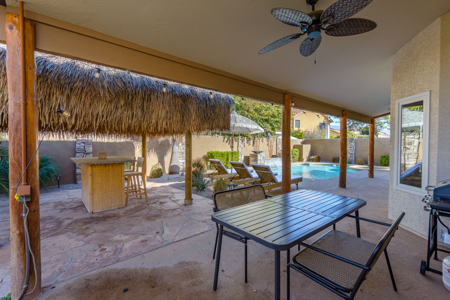 Covered patio dining area with BBQ grill, pool views, and easy access to the tiki lounge and backyard seating.