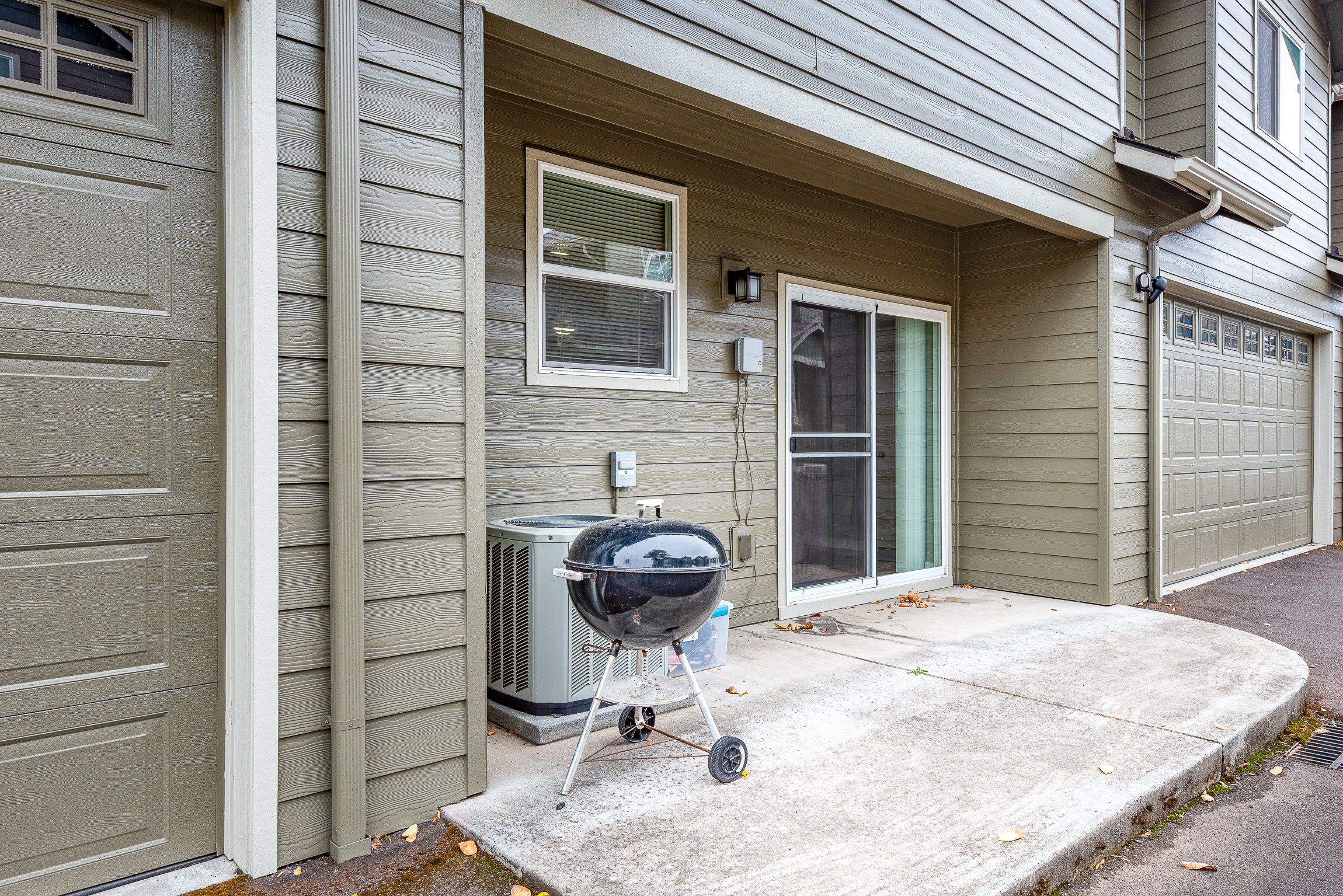 A gray house exterior features a black charcoal grill on a concrete patio next to a central air conditioning unit and a large window with a sliding door, with a garage door visible on the right.