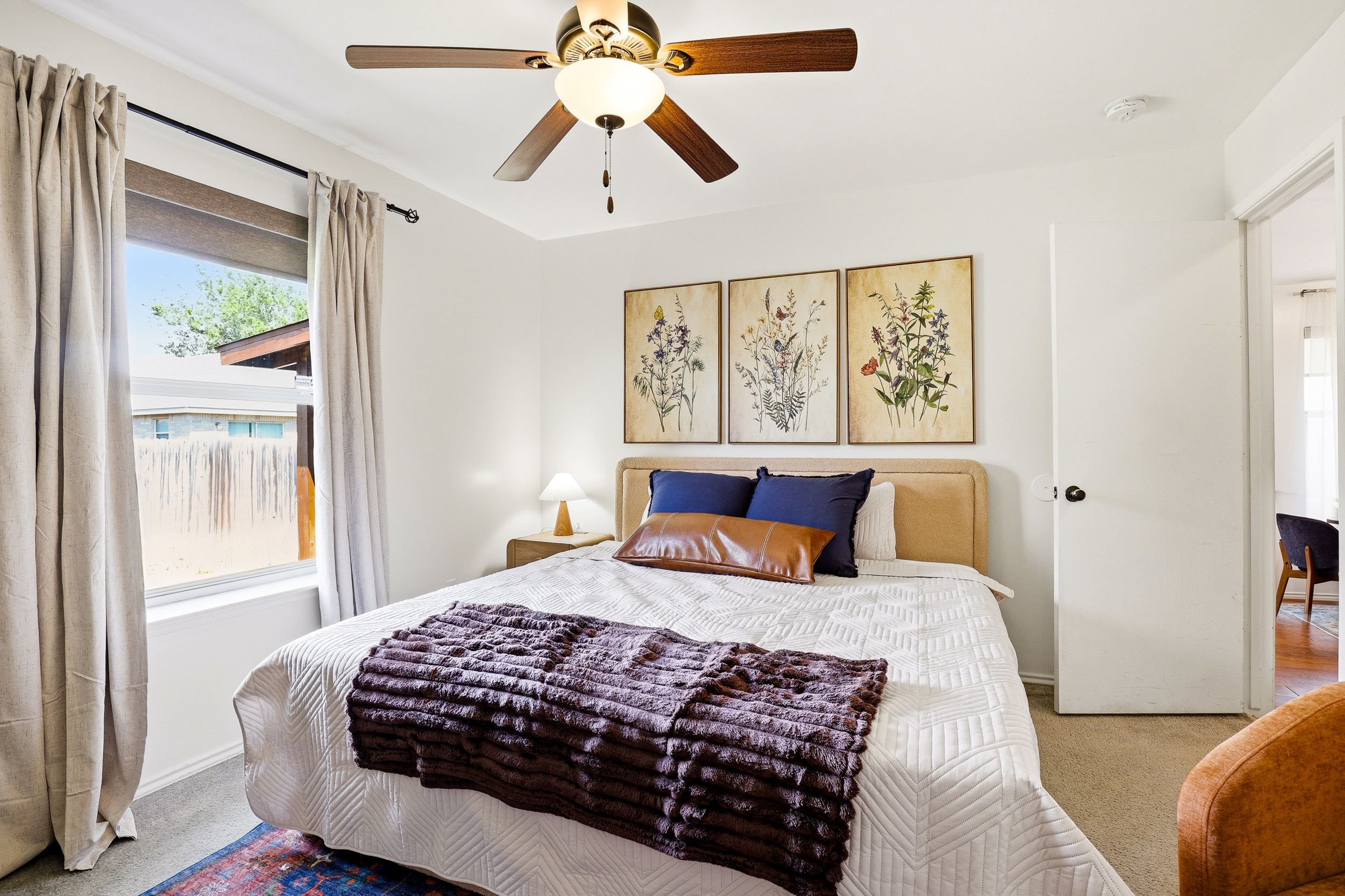Primary bedroom with king bed, botanical wall art, plush bedding, ceiling fan, and closet. Quiet and comfortable with natural light and room-darkening shades for a restful night's sleep.