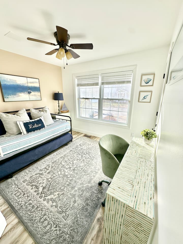 This functional nook features a textured white console desk with a sage green velvet chair. The space is anchored by a large, gray patterned rug over wood-look flooring and sits adjacent to a daybed with a pull-out trundle.