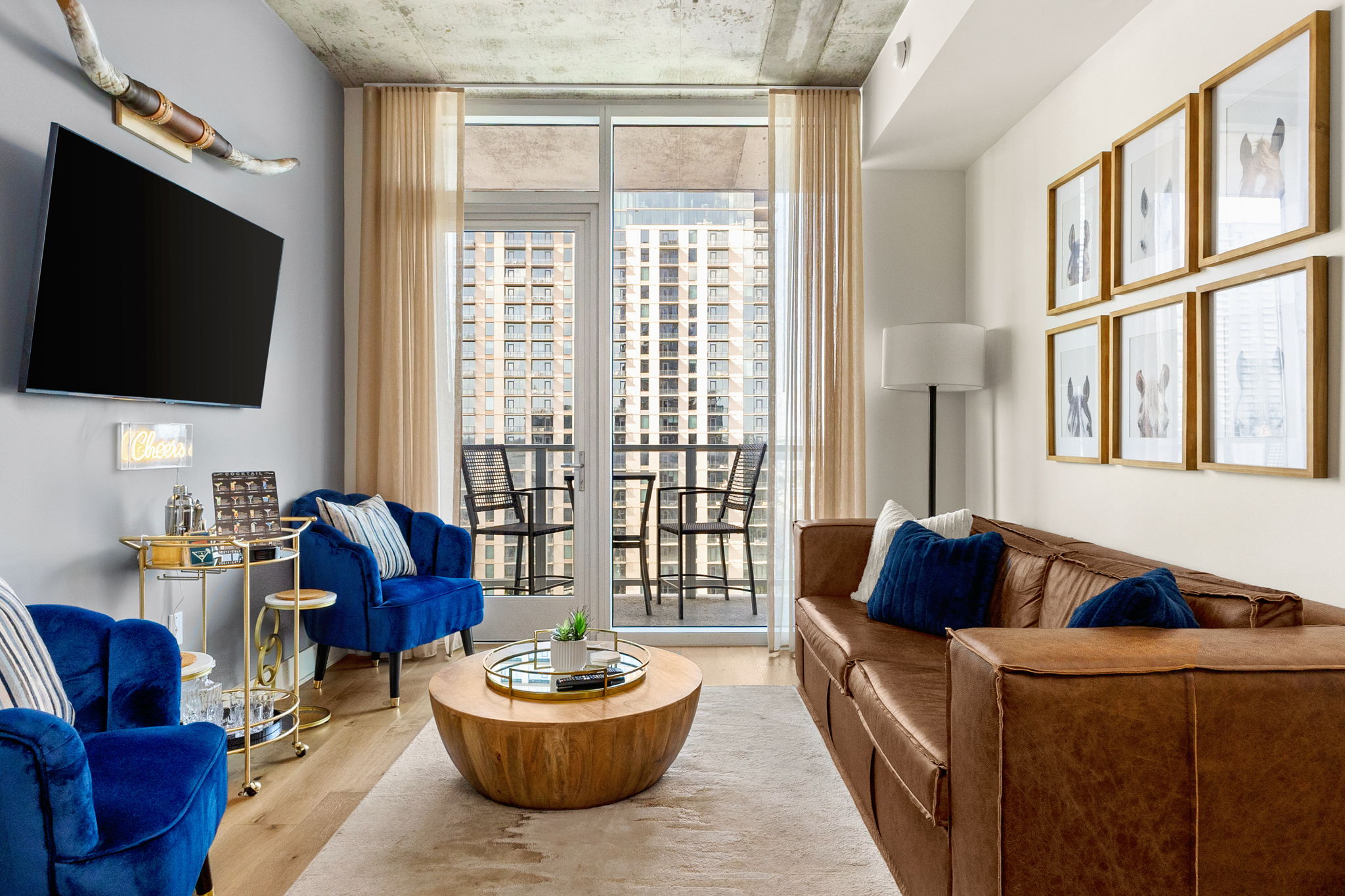 A bright and modern living room featuring a brown leather sofa, two stylish blue armchairs, and a balcony with an urban view, all decorated with a gallery wall of horse portraits and a chic bar cart.