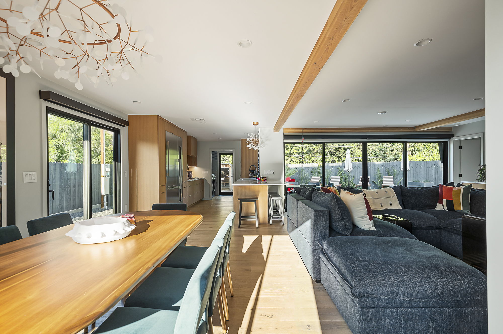 A bright and open living and dining area featuring a long wooden table, a large sectional sofa, and a modern kitchen with light wood cabinetry, all connected by seamless hardwood flooring and exposed beams.