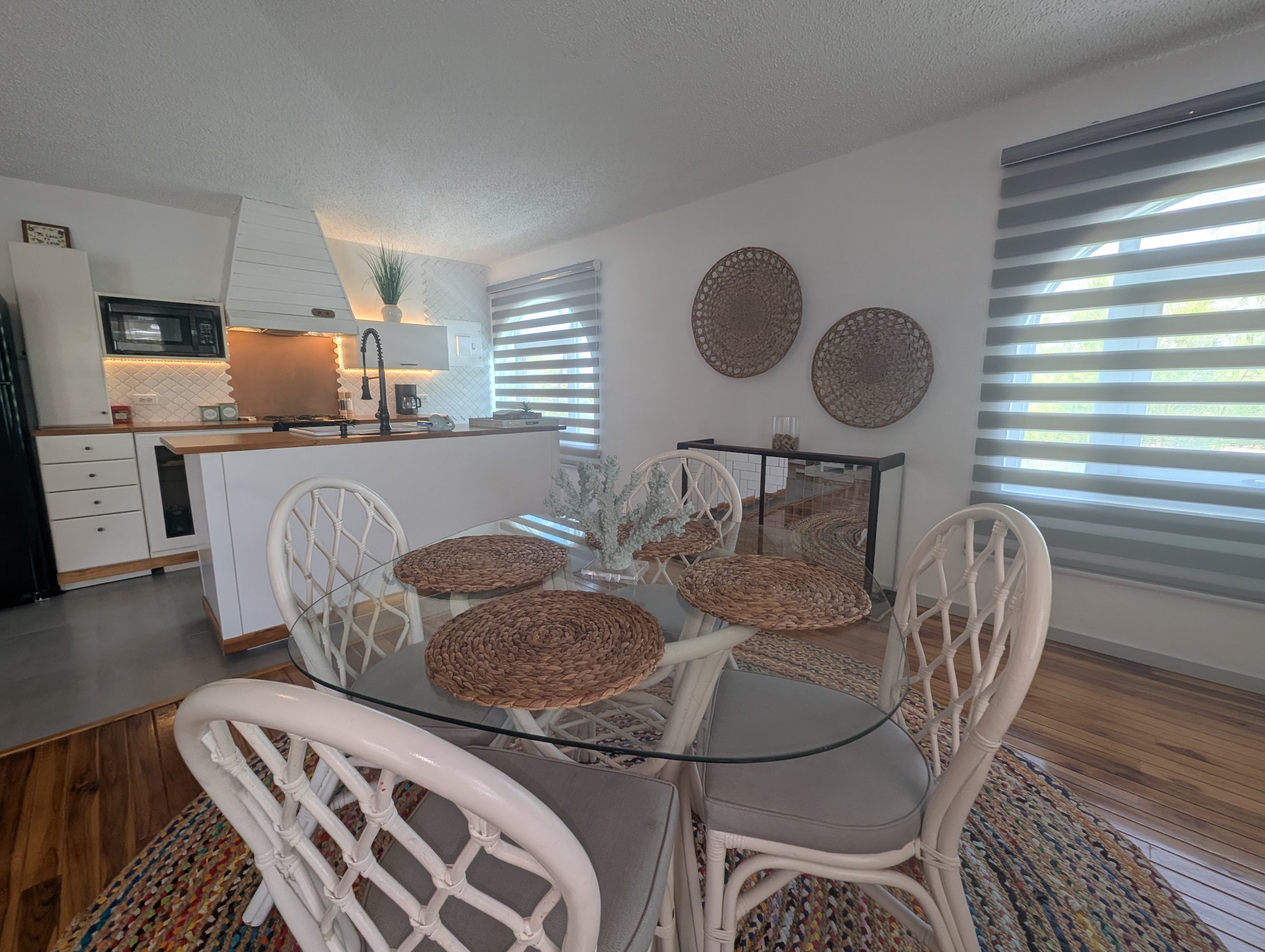 Upstairs dining area at Villa Coral featuring a round glass-top table, white rattan chairs, woven placemats, a colorful jute rug, and a clear view of the adjacent kitchen island in Dorado, Puerto Rico.

