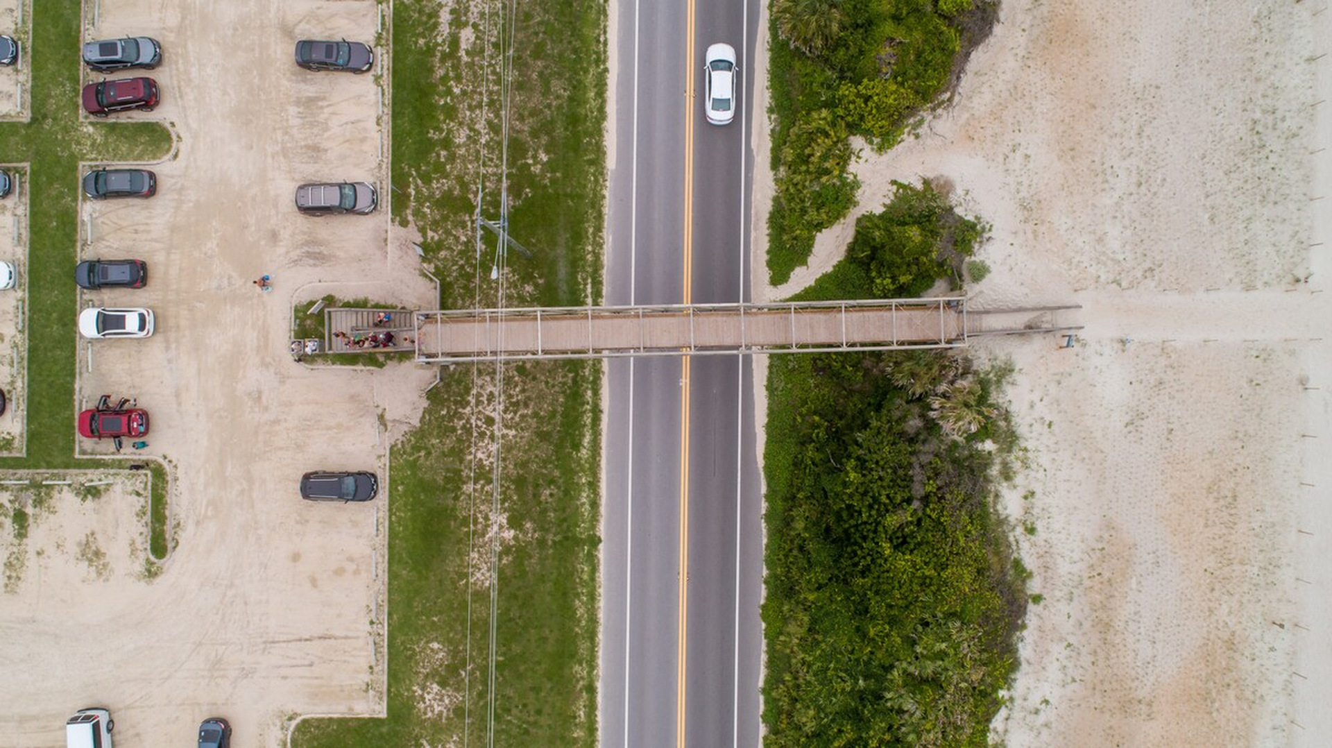 The walkway seen from above, with the car park on one side and dunes and open sand on the other. Roughly eight to ten minutes on foot from the front door, chairs and boogie boards rattling in the wagon behind you.