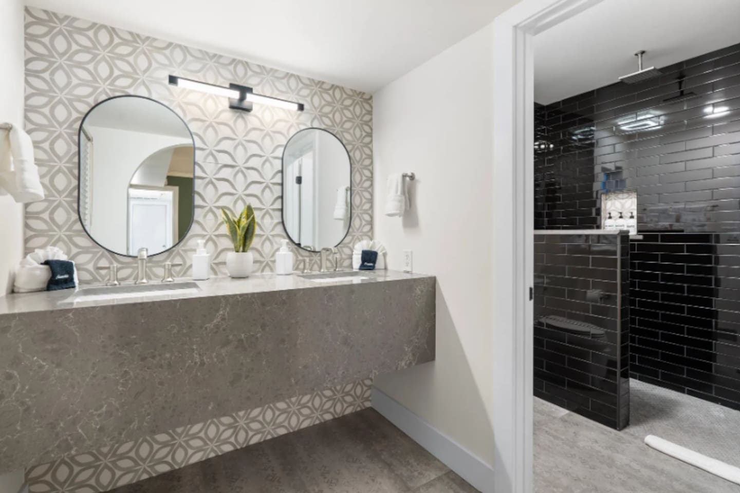 A dual vanity in patterned stone tile sets a spa-like tone, with a moody black-tiled shower just beyond. Every surface here feels intentional, right down to the oval mirrors and brushed fixtures.
