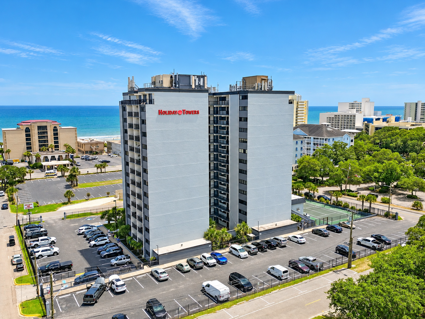 Aerial view of Holiday Towers with shared pool, hot tub, sports court, parking, and Myrtle Beach attractions nearby.