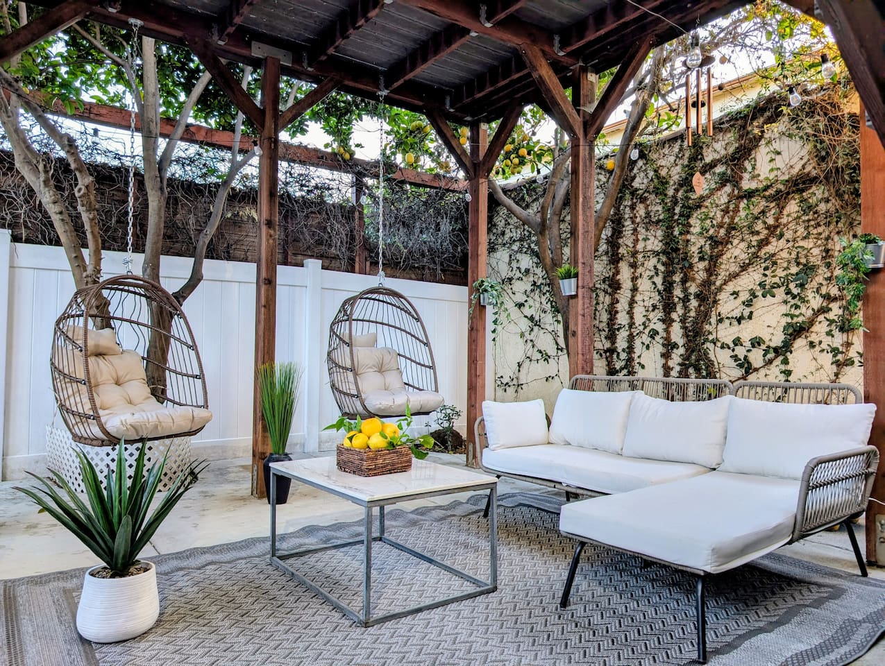 This private, gated, and completely secluded covered patio offers total peace of mind with handy driveway access. A rustic, solid-roofed wooden pergola shelters a grill, sleek sectional, plush cushions, and twin hanging teardrop swings.
