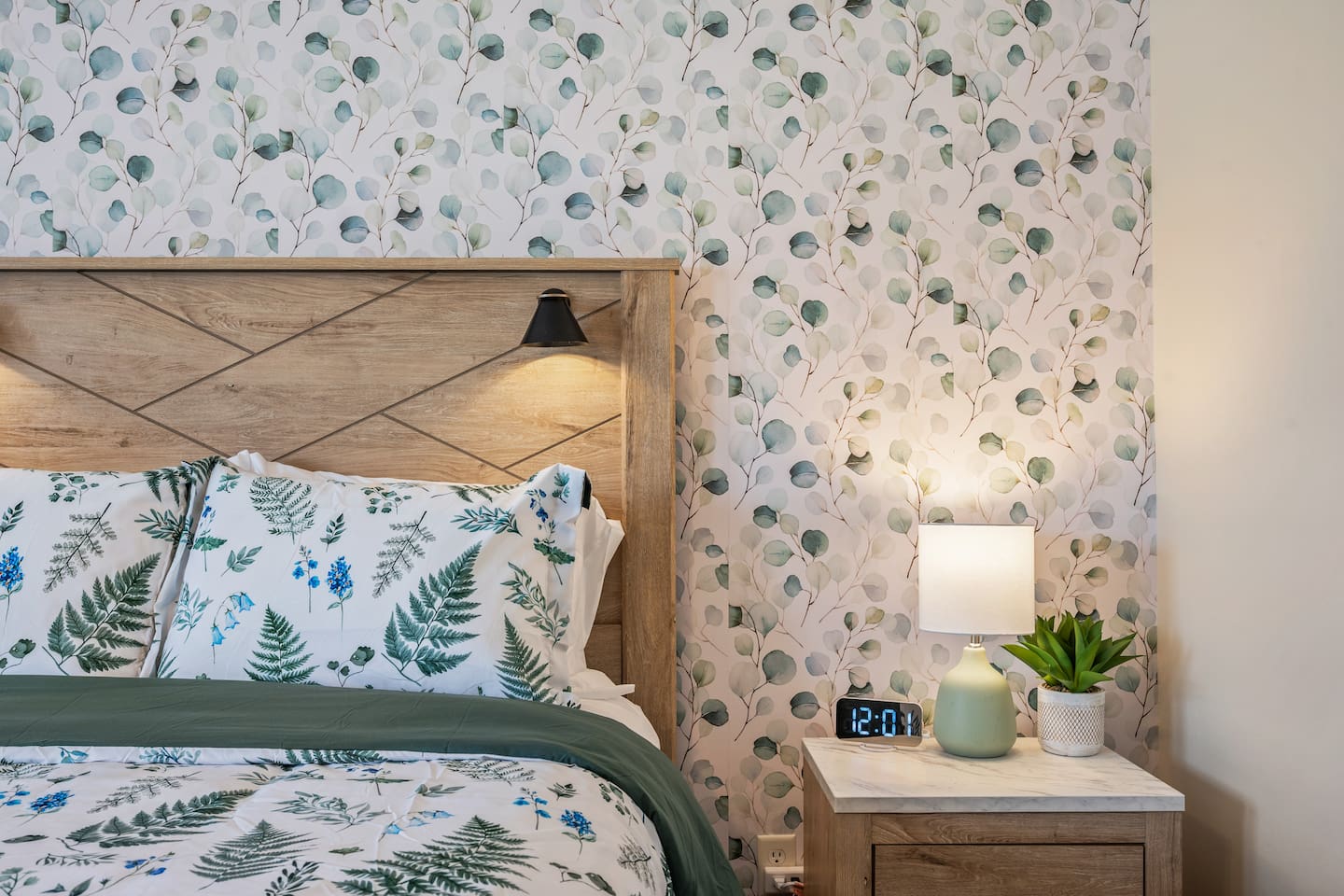 This serene bedroom features cozy botanical bedding, soft lighting, and a stylish wood headboard. Thoughtful touches like a bedside lamp, digital clock, and greenery add to the calm, restful vibe. 🌿🛏️💤