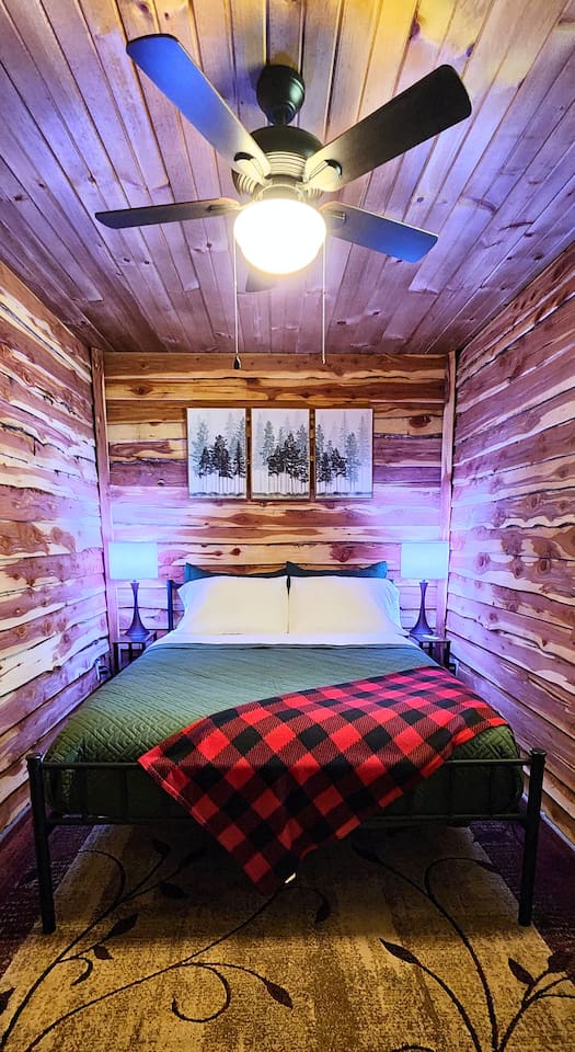 Rest easy in this inviting queen bedroom featuring cedar-lined walls, soft lighting, and forest-inspired decor. A classic buffalo plaid throw and cozy quilt add warmth, while a ceiling fan overhead keeps the room fresh and relaxing all night.
